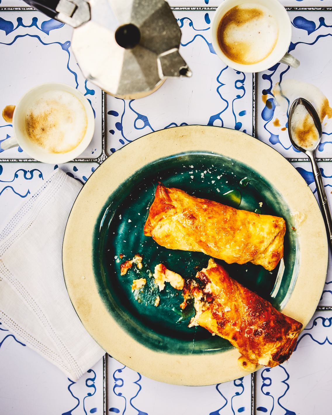 Two quesito pastries on a green plate with a coffee setup on a patterned tablecloth.